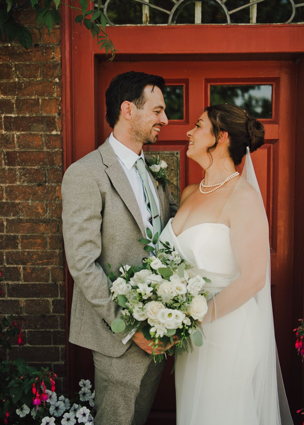 A bride and groom smile at each other in front of a red door, she holds a white bouquet and wears a veil and pearl necklace.