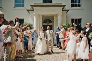 Bride and groom exit wedding venue through confetti shower, surrounded by cheering guests outside elegant white manor.