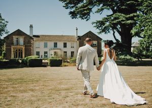 Bride and groom walking hand in hand toward an elegant country house venue on a sunny wedding day.