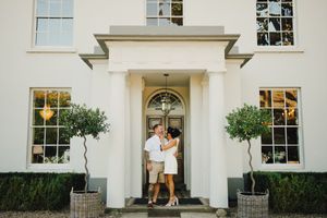 Couple embracing at the entrance of an elegant white manor house with topiary trees and a grand front door.
