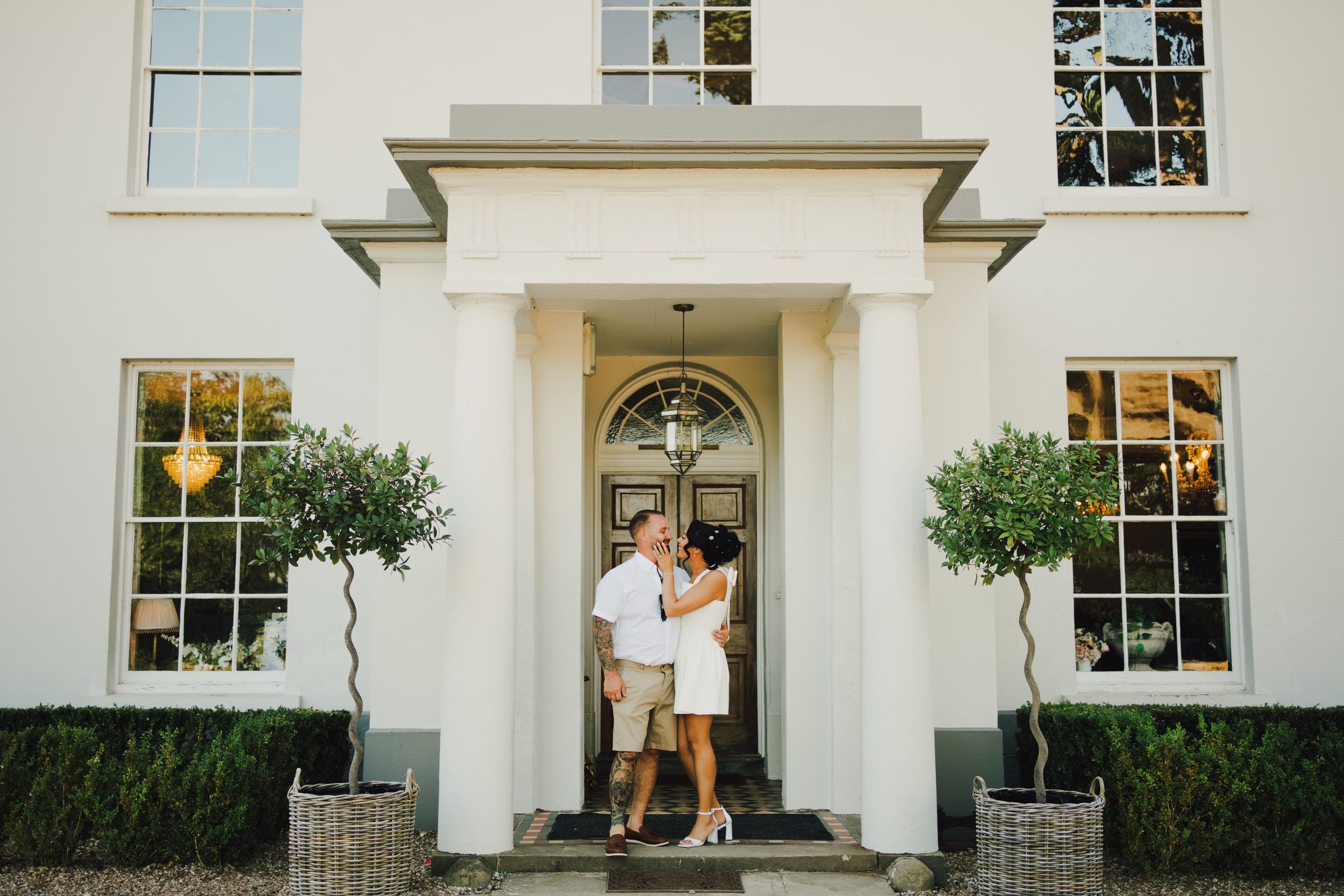 Couple embracing at the entrance of an elegant white manor house with topiary trees and a grand front door.
