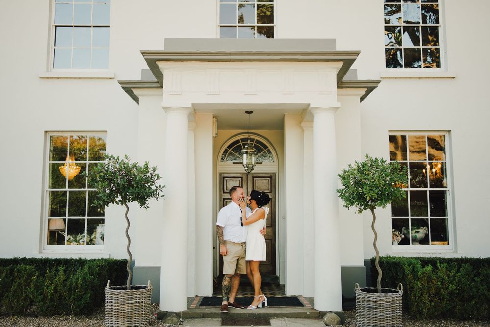 Couple embracing at the entrance of an elegant white manor house with topiary trees and a grand front door.