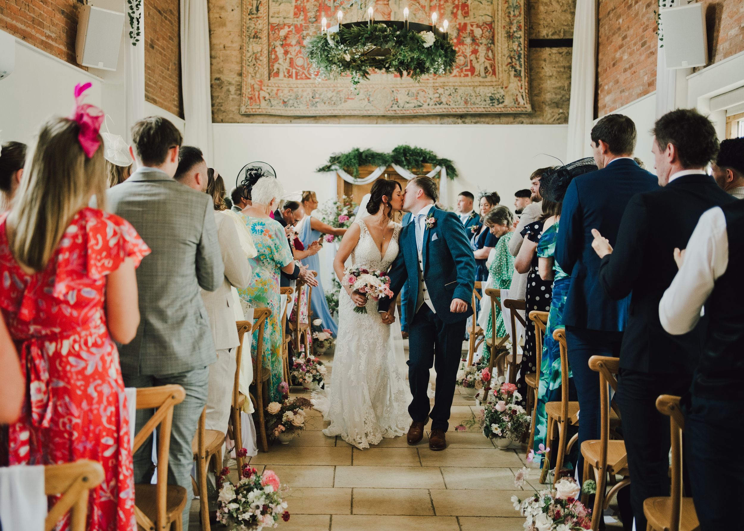 Bride and groom walk down the aisle at an elegant indoor wedding ceremony with floral decorations.