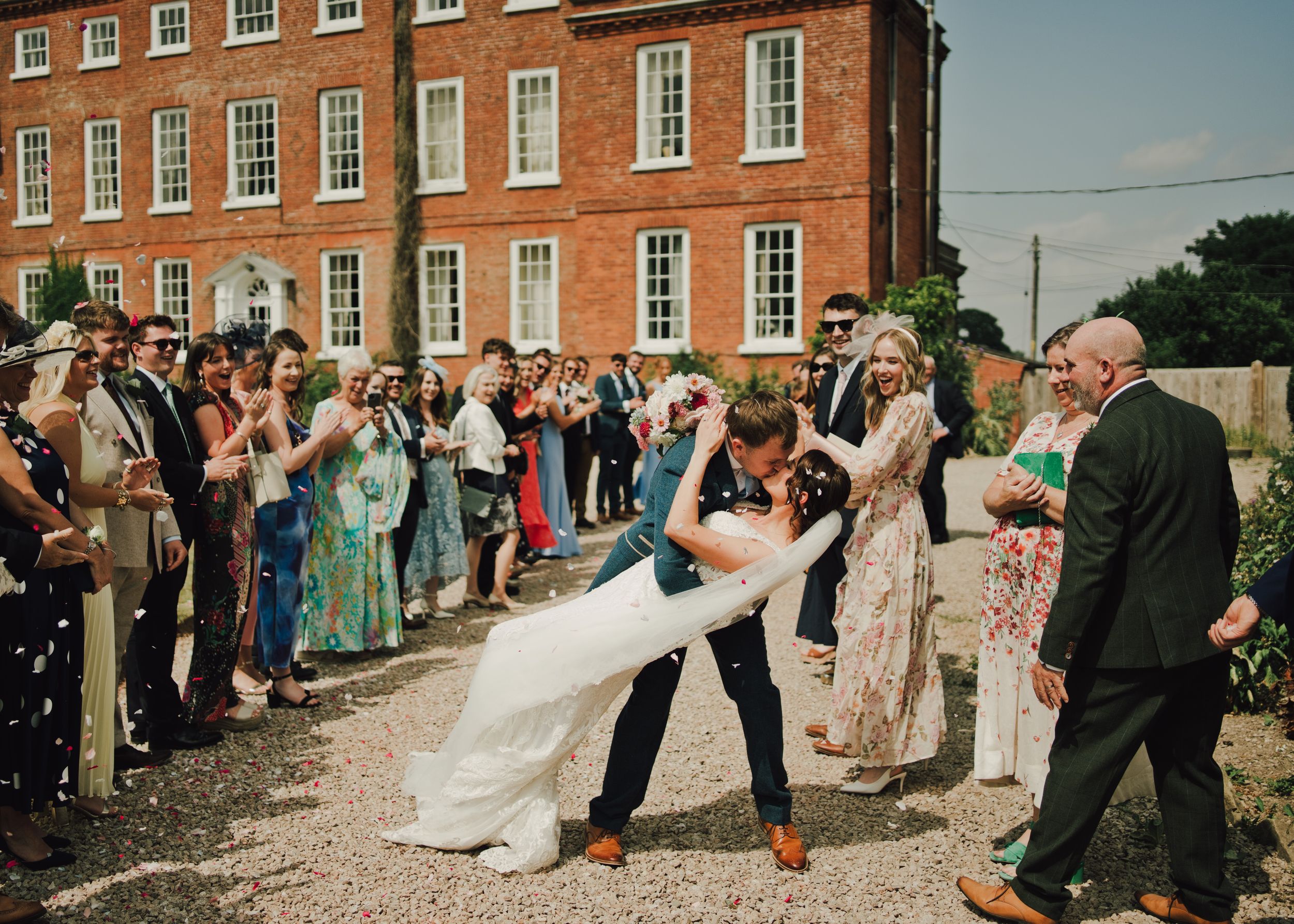 Groom dips bride in a romantic dance outside a red brick manor, surrounded by cheering wedding guests.