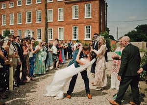 Groom dips bride in a romantic dance outside a red brick manor, surrounded by cheering wedding guests.