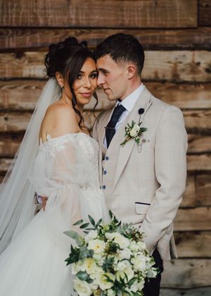 Romantic wedding couple in white attire share intimate moments against rustic wooden wall backdrop with floral bouquet.