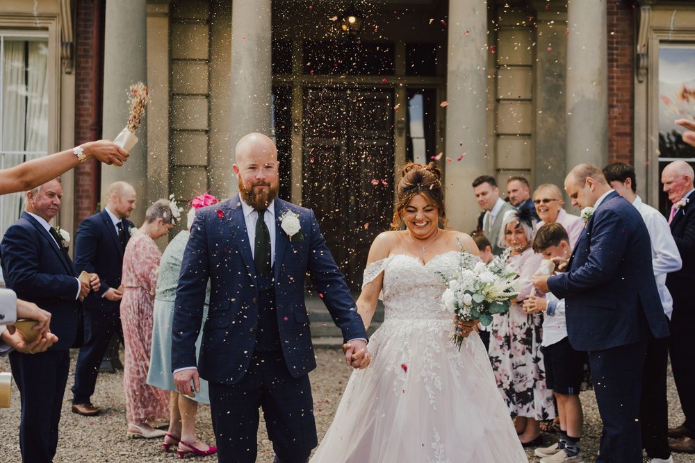 Wedding couple exits historic building while guests shower them with confetti during celebratory moment.
