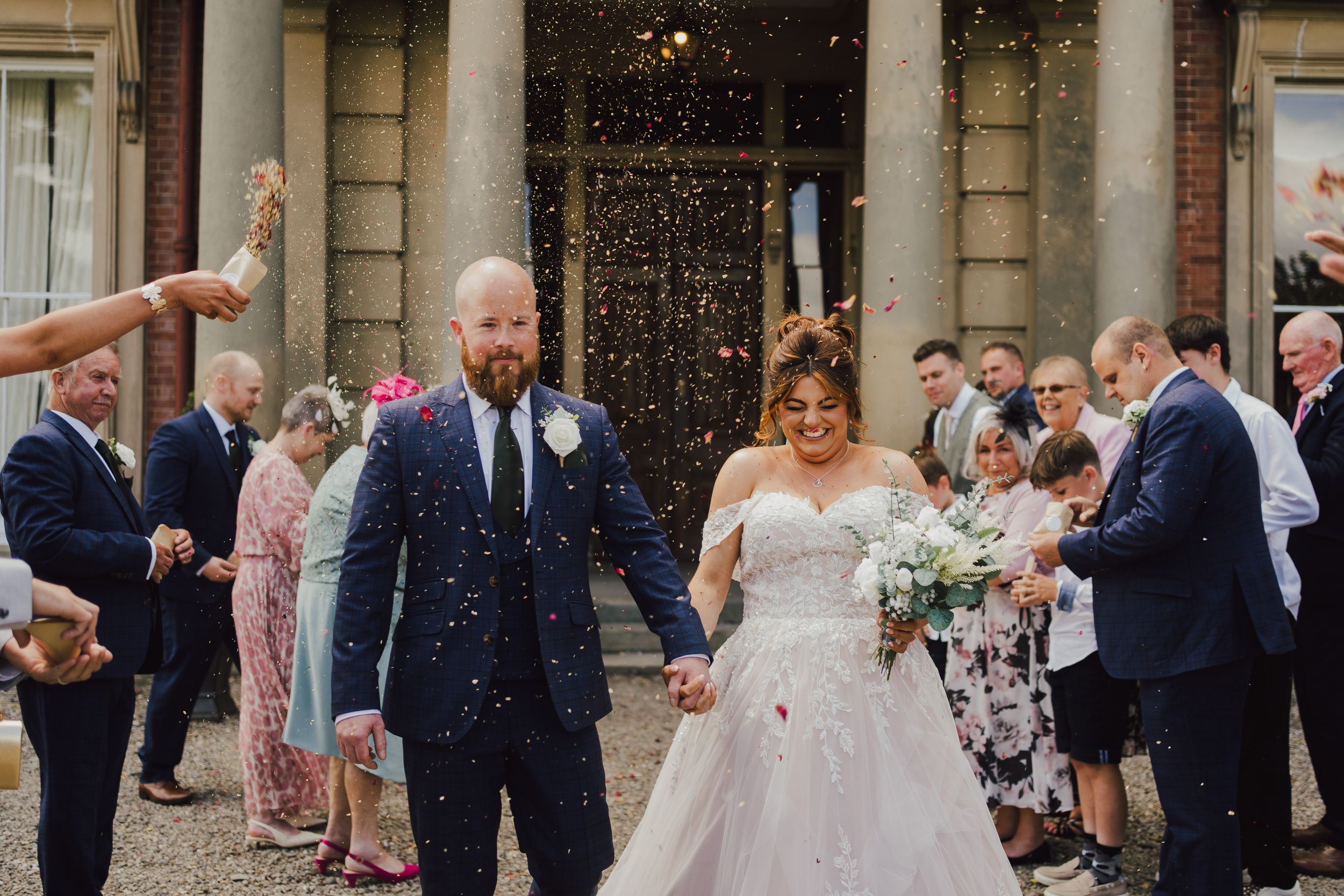 Wedding couple exits historic building while guests shower them with confetti during celebratory moment.
