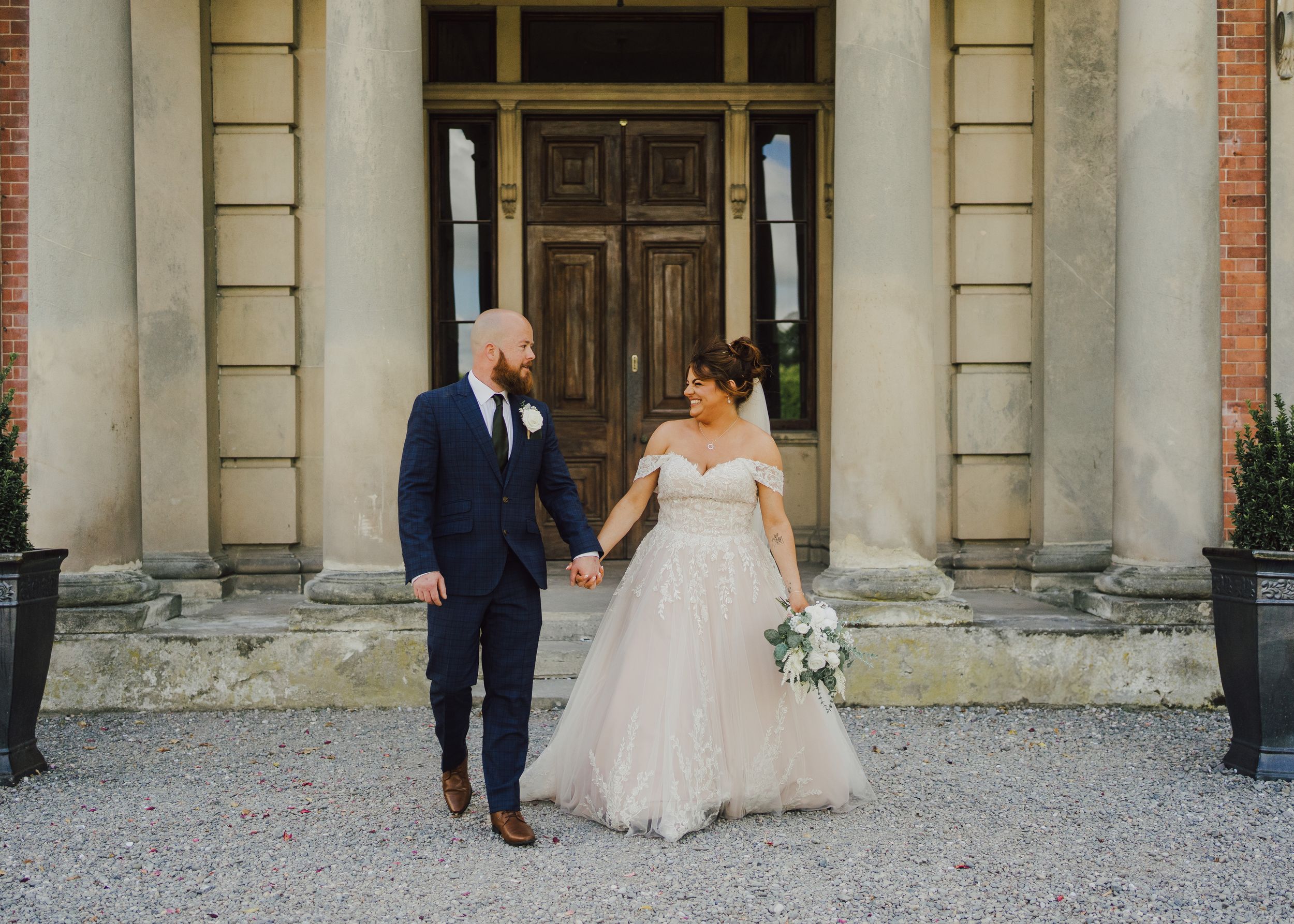 A couple holding hands on their wedding day stand in front of a grand entrance with columns and wooden doors.