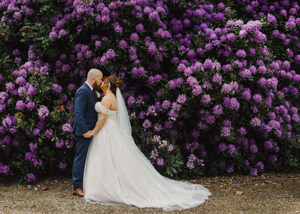 A couple shares a romantic embrace in front of a vibrant wall of purple lilac blooms on their wedding day.