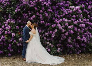 A couple shares a romantic embrace in front of a vibrant wall of purple lilac blooms on their wedding day.