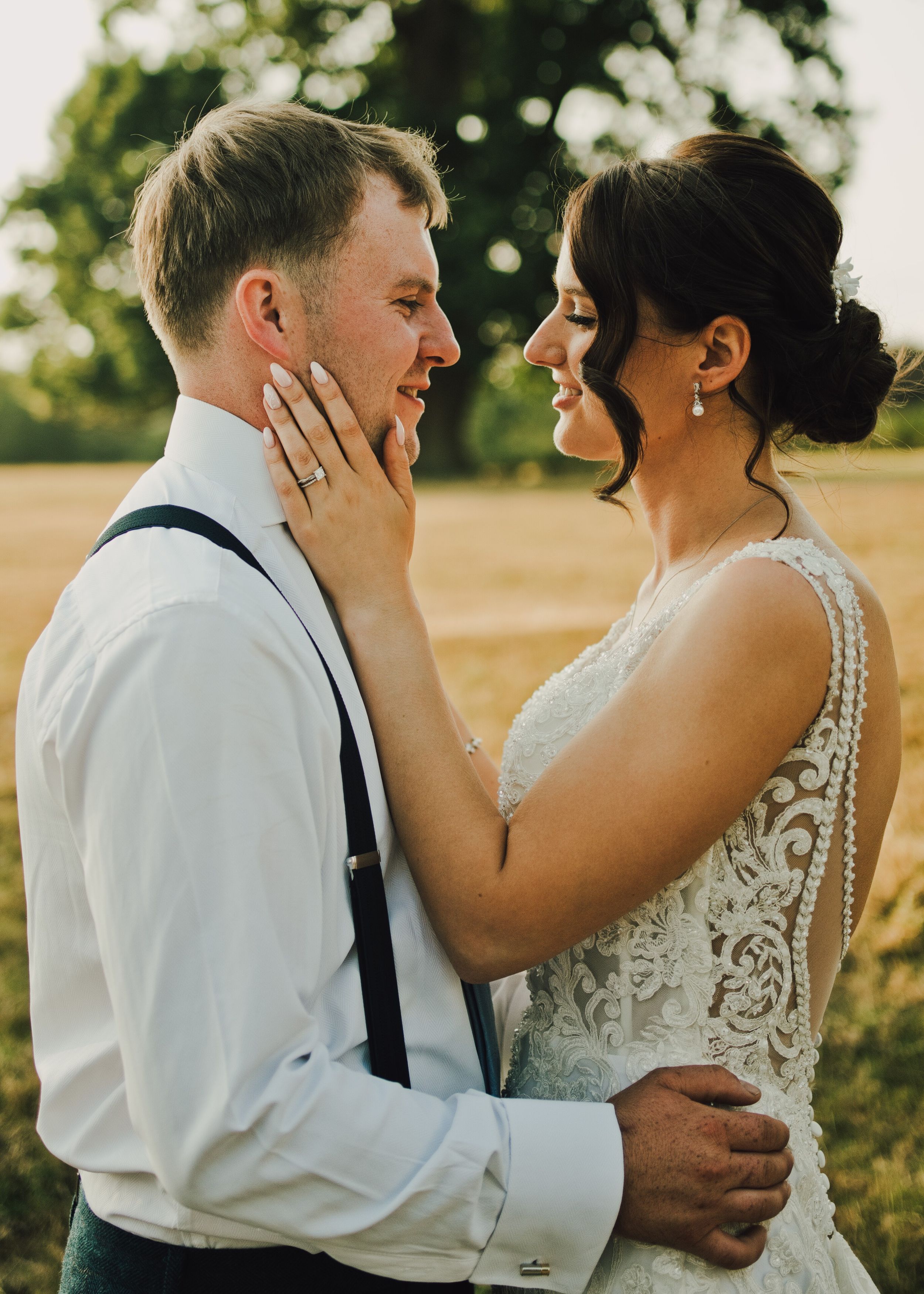 A bride and groom share a romantic moment in a sunlit field, with the bride gently touching the grooms face.