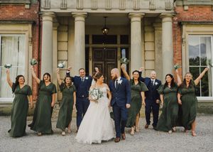 Wedding party guests gather for a panoramic group photo outside a historic brick mansion.