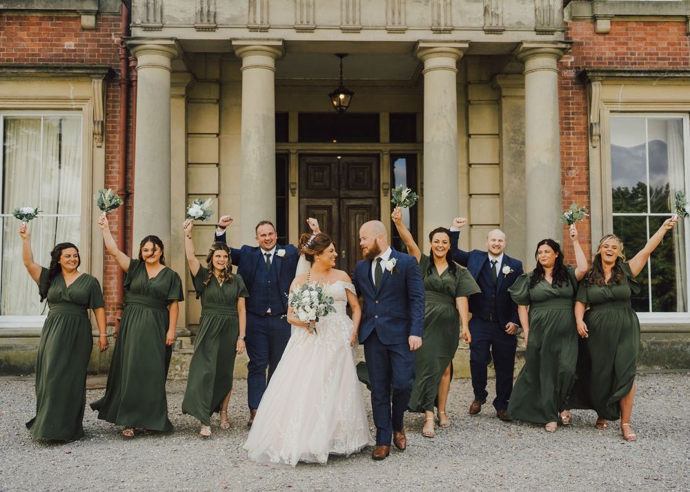 Wedding party guests gather for a panoramic group photo outside a historic brick mansion.