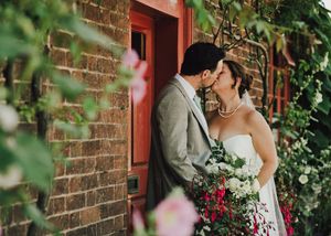 Bride and groom sharing a kiss outside a brick wall with red door, surrounded by lush greenery and flowers.