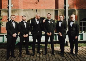 Six groomsmen in black tuxedos with bow ties posing together outside a brick building at a wedding.