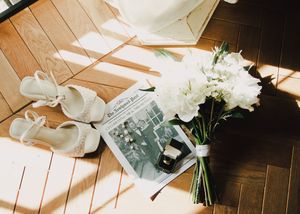 White bridal shoes, a newspaper, and a white floral bouquet arranged on a wooden floor in soft sunlight.