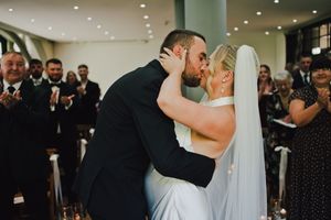 Bride and groom share their first kiss at wedding ceremony while guests applaud in elegant venue.