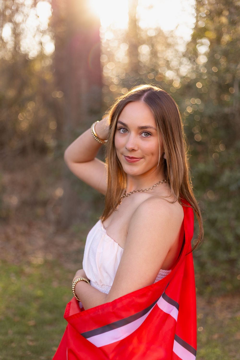 Young woman in white dress wrapped in a red blanket posing in a sunlit forest at golden hour.