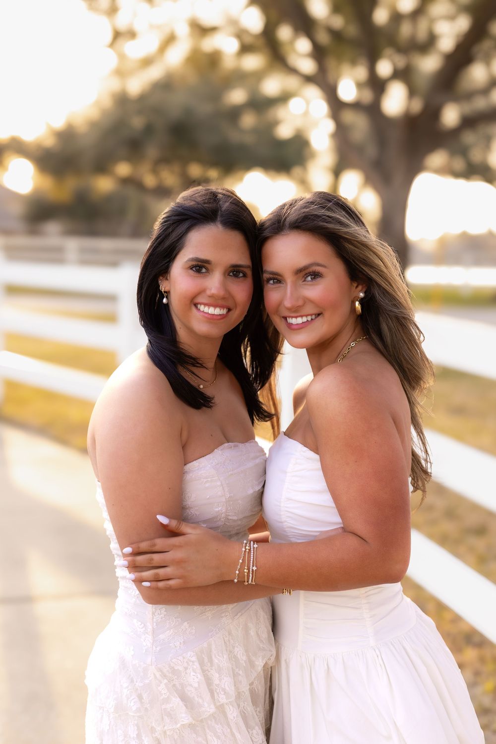 Two smiling young women in matching white strapless dresses embracing near a white fence at golden hour outdoors.