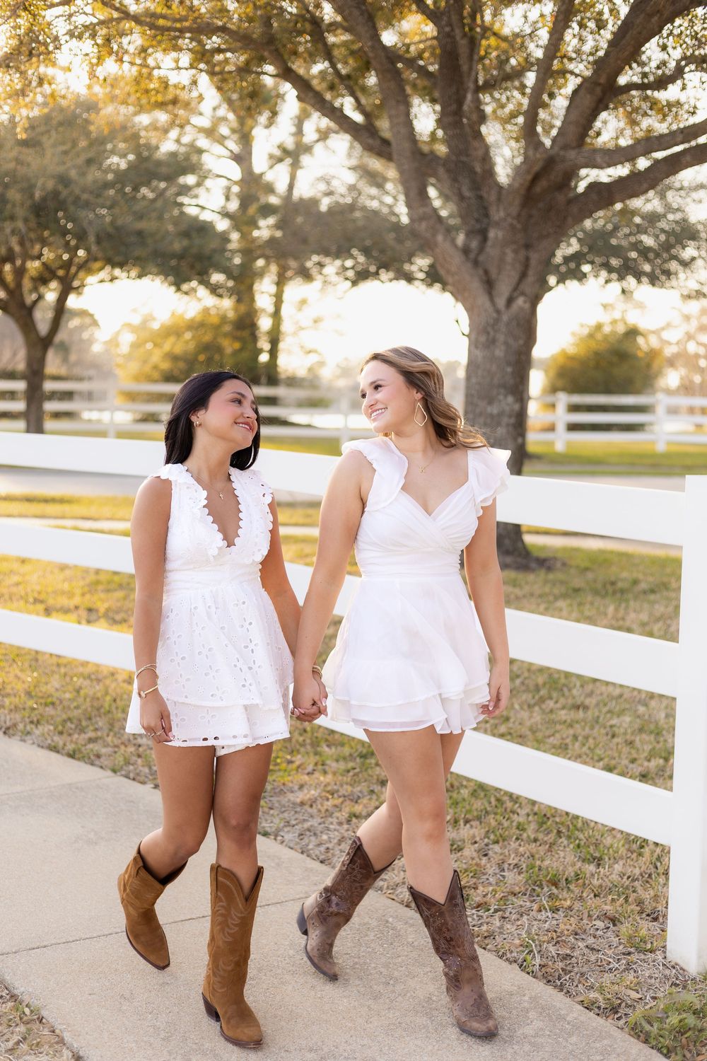 Two young women in white dresses and cowboy boots hold hands while walking along a white fence at golden hour.