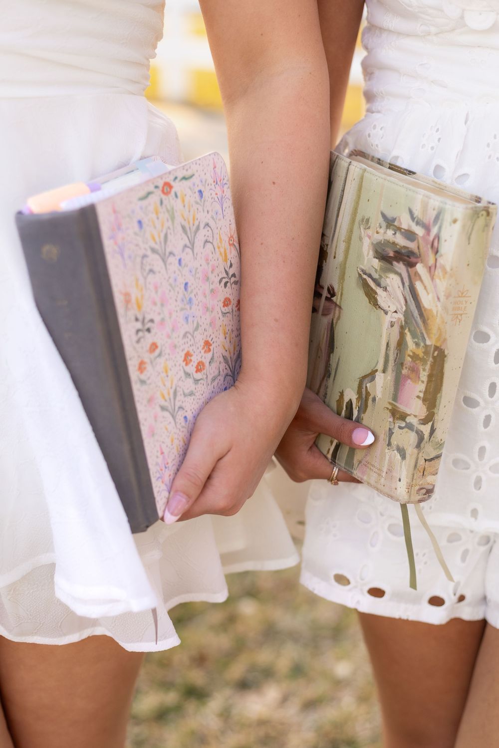 Close-up of two women in white dresses holding decorative floral-covered journals outdoors.