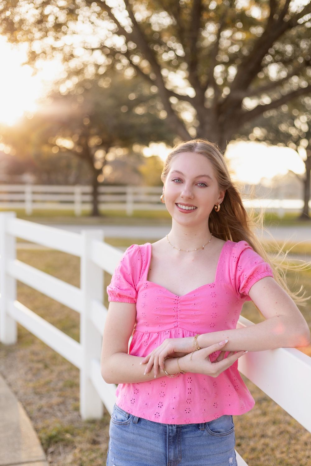 Blonde teen girl in pink top and jeans smiling at white fence during golden hour sunset.