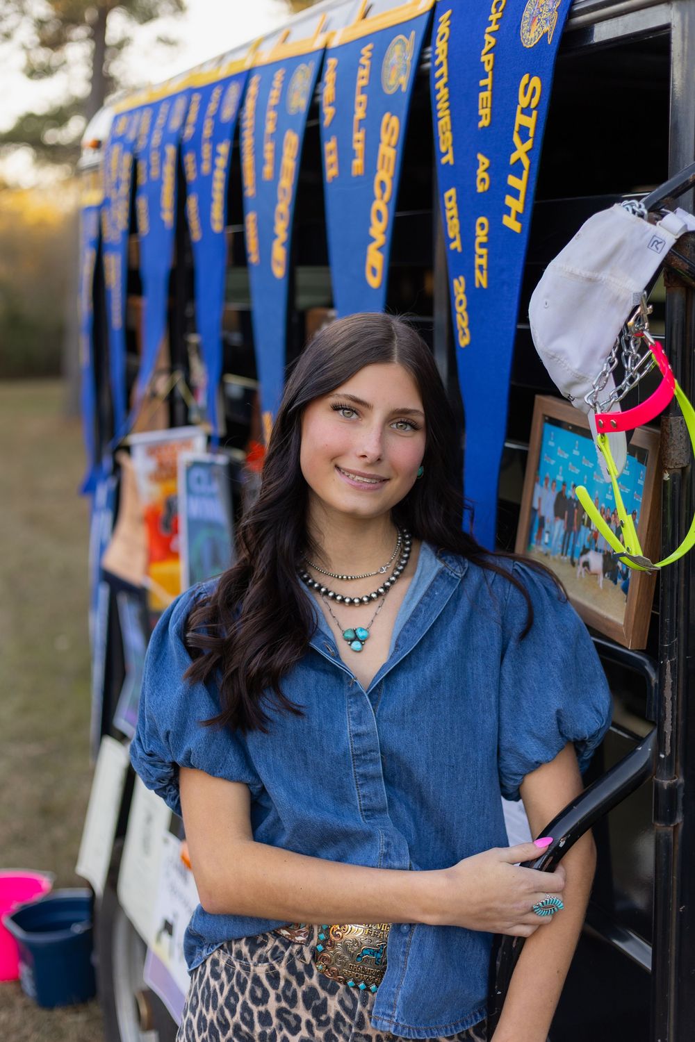 Smiling brunette woman in denim top with turquoise jewelry posing by trailer with blue FFA award banners.