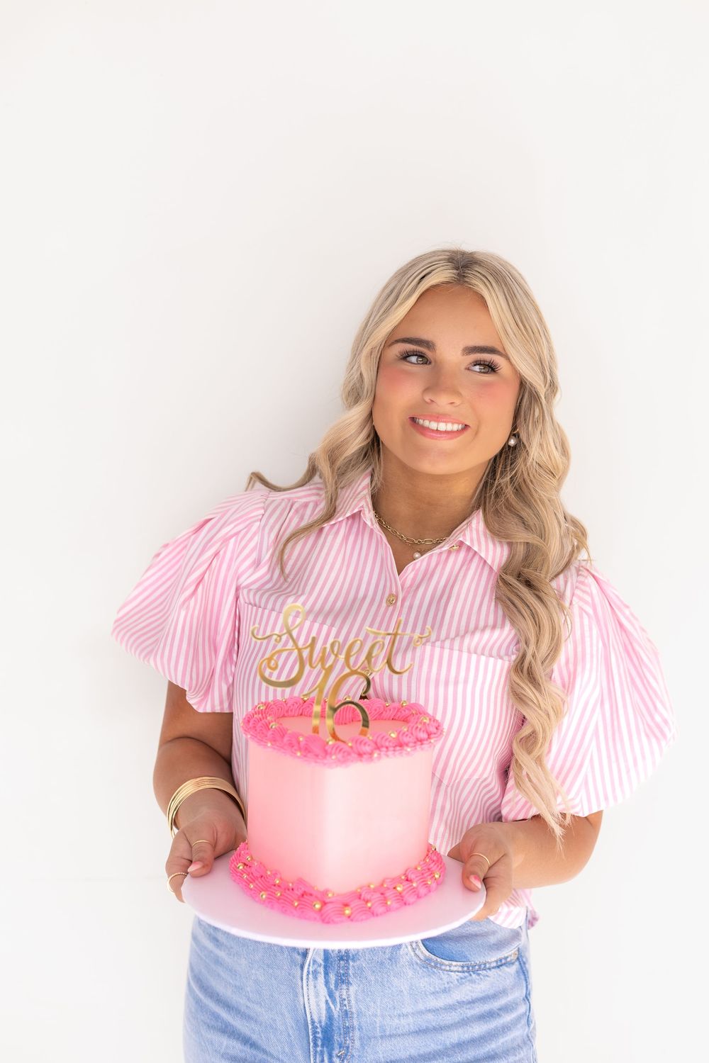Smiling blonde teen holding a pink Sweet 16 birthday cake with gold topper against white background.