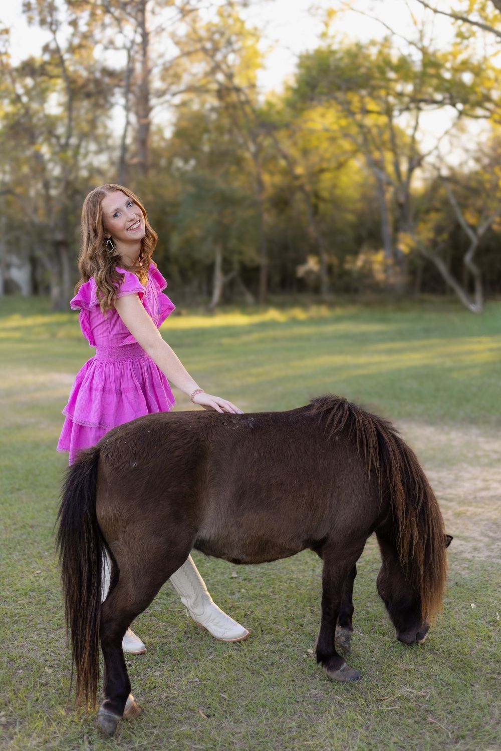 Young woman in pink dress and white boots smiling while petting a dark miniature horse outdoors.
