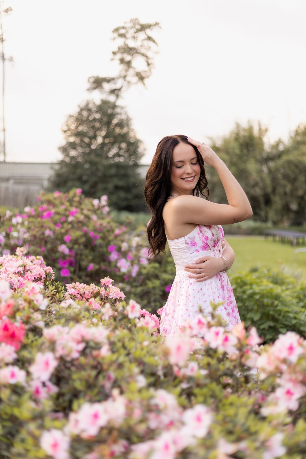 Brunette woman in floral pink dress smiling among blooming pink azalea bushes in garden.