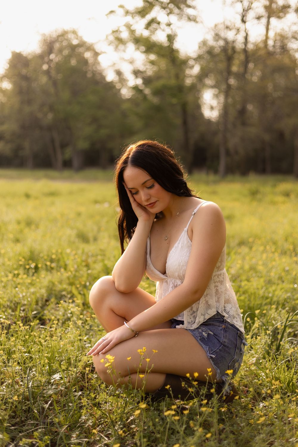 Young brunette woman in white lace top and denim shorts sitting cross-legged in sunny meadow.