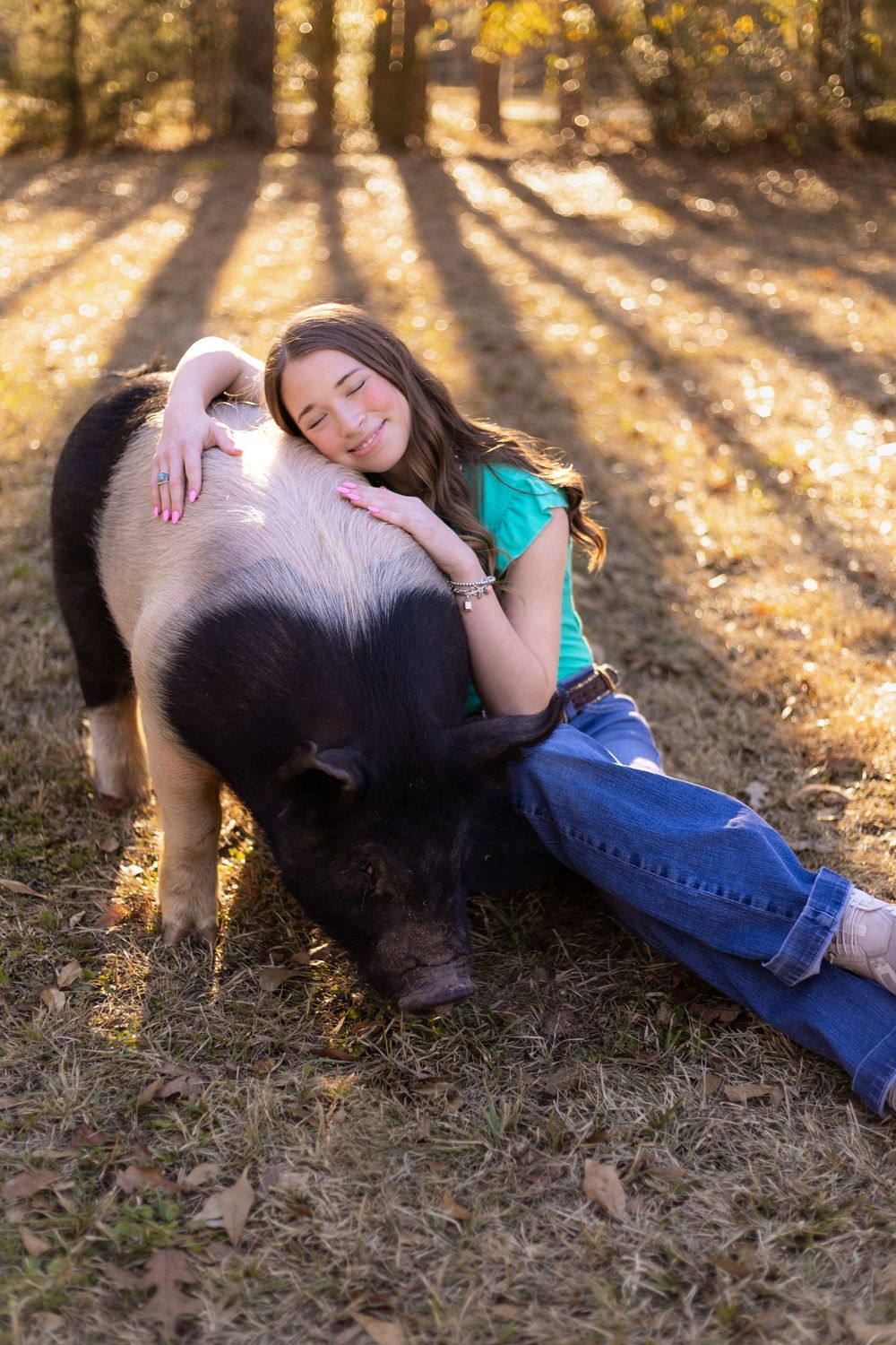 Smiling girl in teal top and jeans cuddling with a large black and white pig on grass.