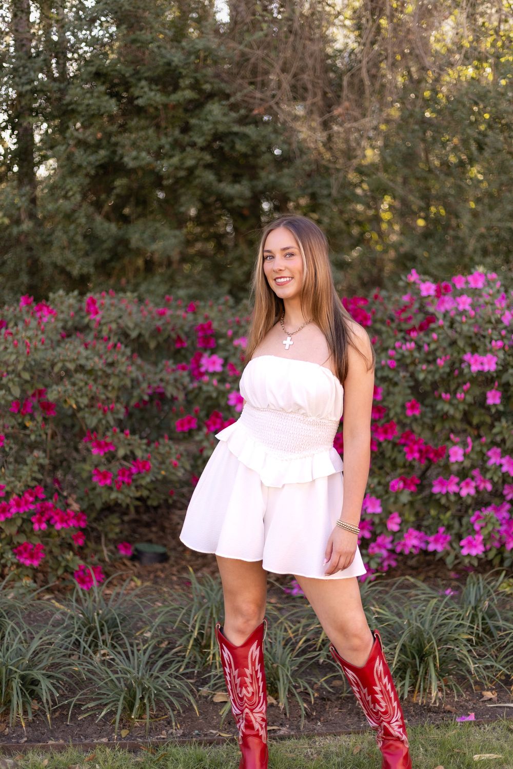 Smiling young woman in white peplum dress and red cowboy boots posing before vibrant pink azalea bushes outdoors.