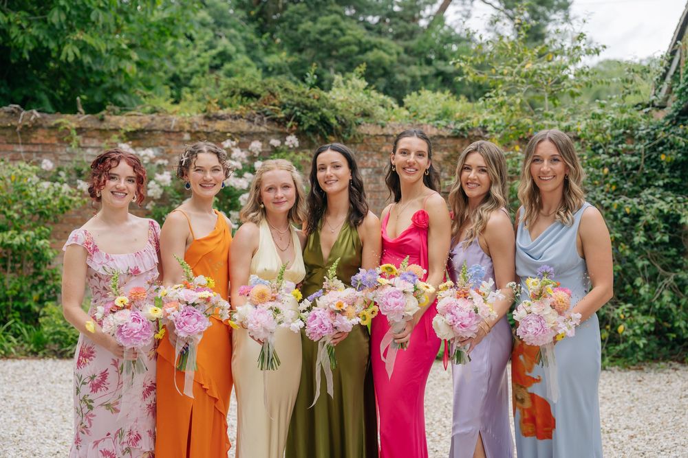 Seven bridesmaids in colorful dresses holding pink bouquets pose outdoors at a garden wedding venue. cotswolds wedding photographer