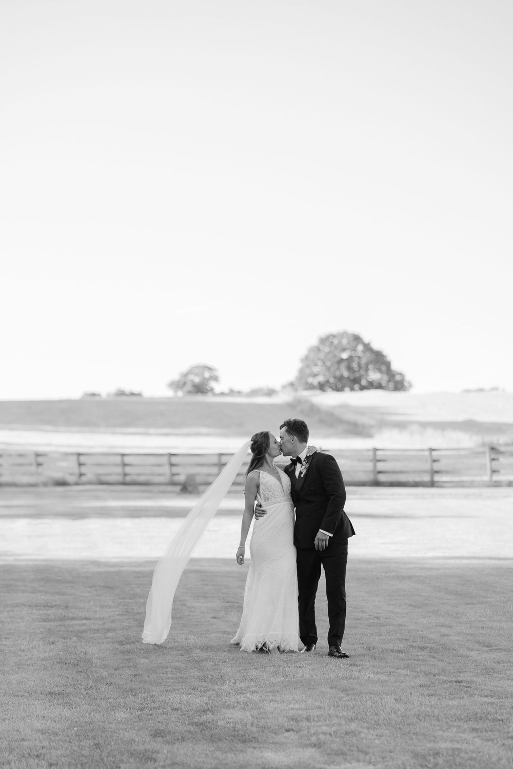 Black and white photo of a bride and groom sharing a kiss outdoors on a grassy field with a wooden fence and trees. cotswolds wedding photo