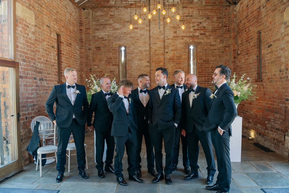 Groomsmen in black tuxedos and bow ties laughing together in a rustic brick venue with warm Edison bulb lighting. cotswolds wedding photogra