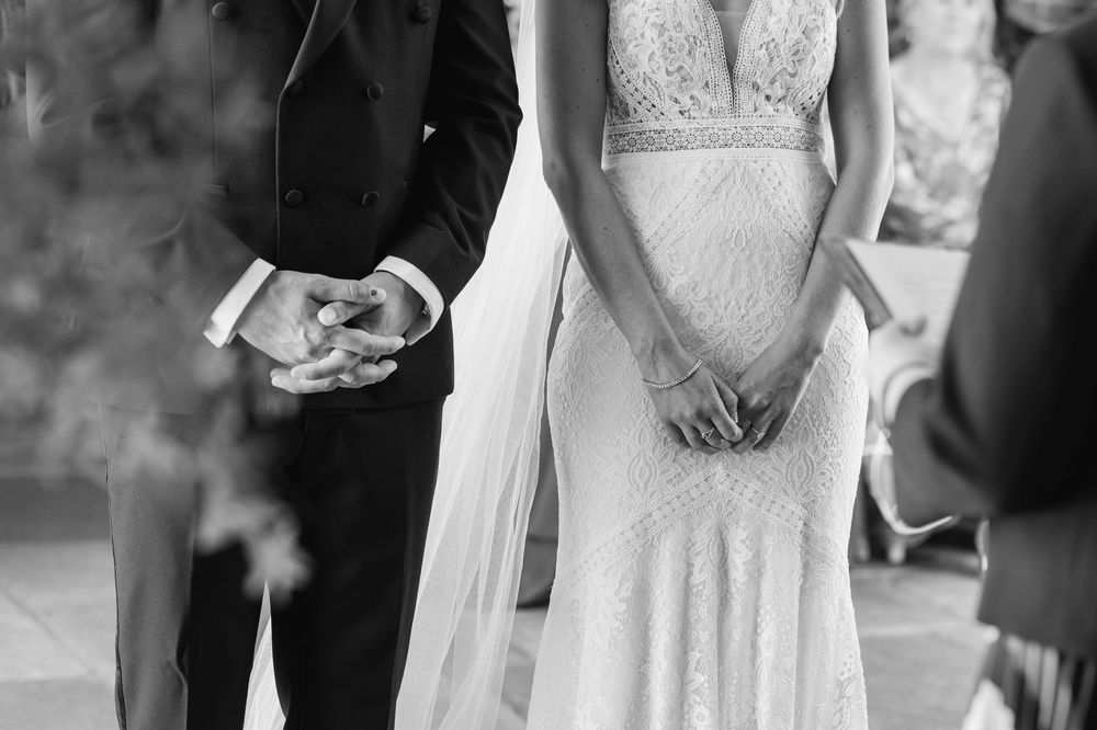 Black and white photo of bride in lace gown and groom in tuxedo standing together during wedding ceremony. cotswolds wedding photographer