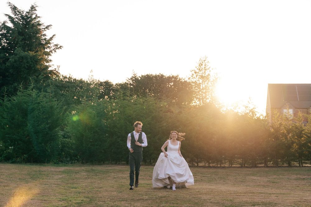Bride and groom walking in golden sunset light on green lawn with trees and barn in background. cotswolds wedding photographer