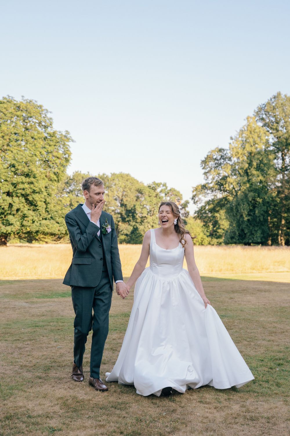 Laughing bride in white ballgown holds hands with groom in dark green suit outdoors on sunny wedding day.
