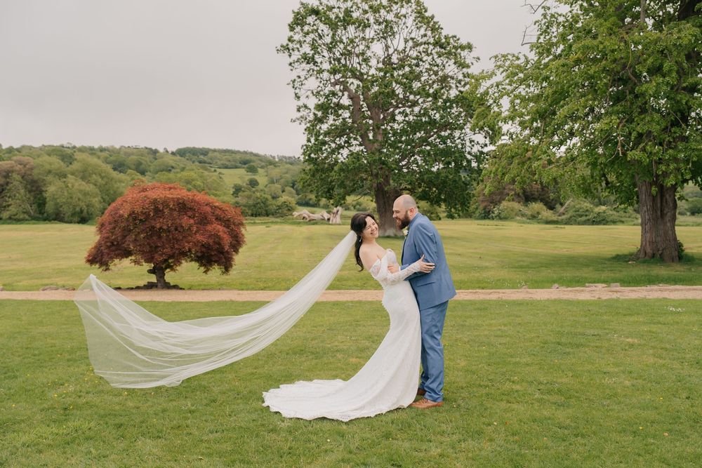 Bride and groom share a romantic moment on lush green grounds, her veil flowing in the wind near a red maple tree.