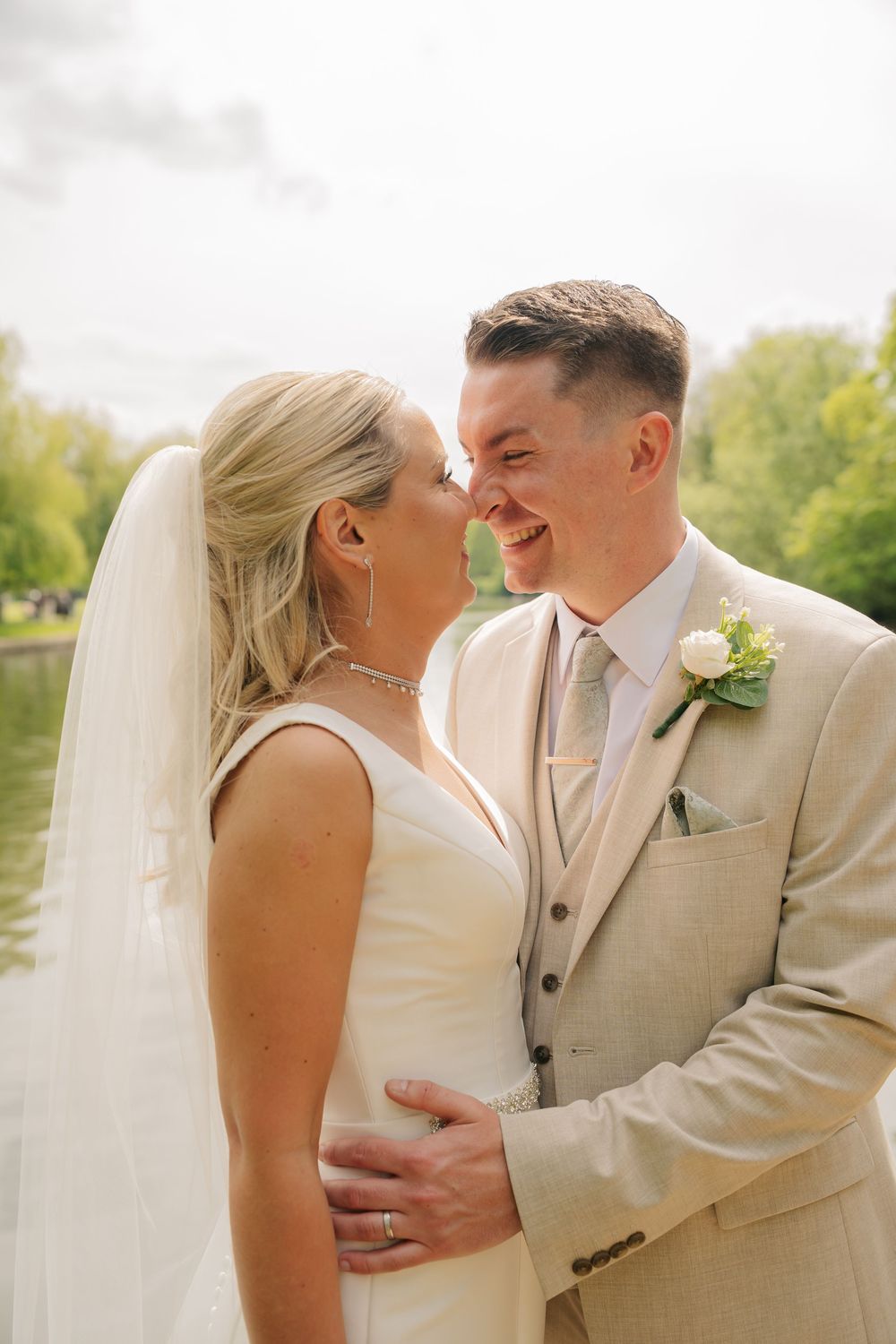 A smiling bride and groom share a romantic moment outdoors near a lake, dressed in a white gown and beige suit.
