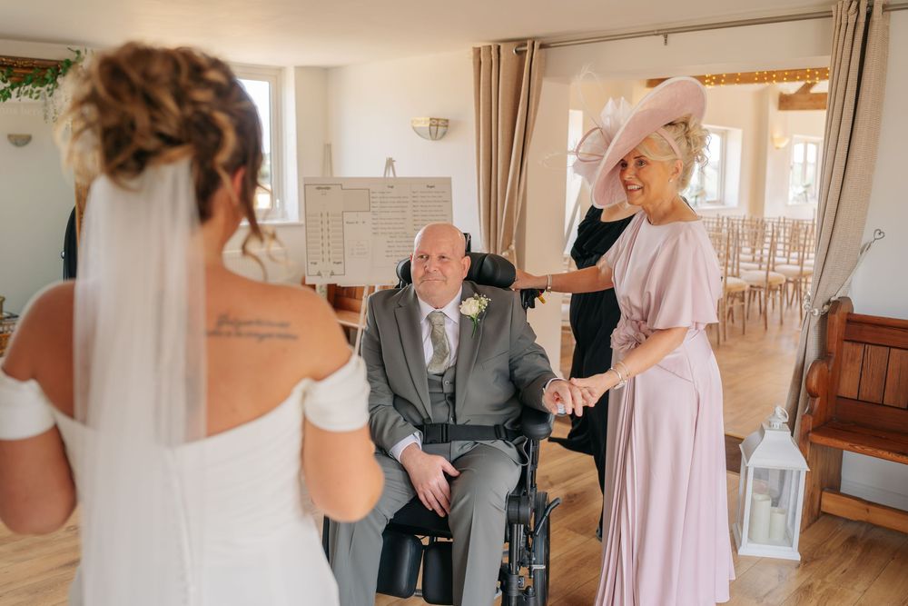 Bride in white gown and veil greets elderly man in wheelchair and woman in pink dress at a wedding venue.