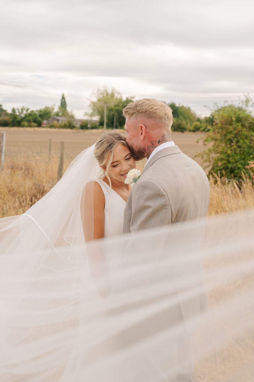 Bride and groom share a romantic moment outdoors, with her veil flowing in the wind against a rustic countryside backdrop.