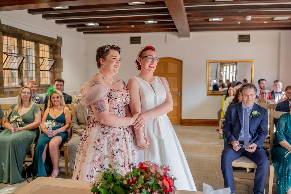 Two brides smiling together during wedding ceremony in rustic venue with seated guests in background. warwickshire wedding photographer