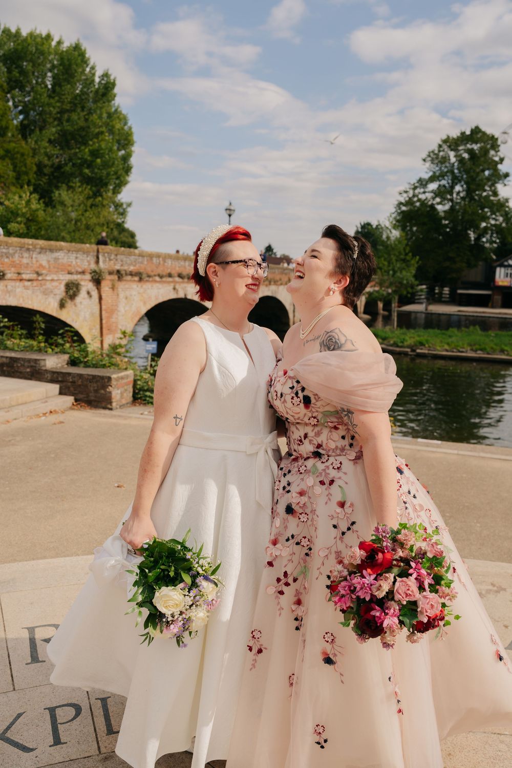 Two brides in wedding dresses holding bouquets laugh together near a river bridge on their wedding day. warwickshire wedding photographer