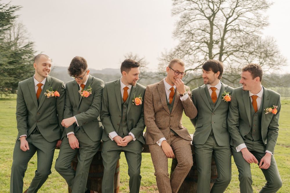 A group of groomsmen in suits strike a playful pose while laughing together in an outdoor setting.