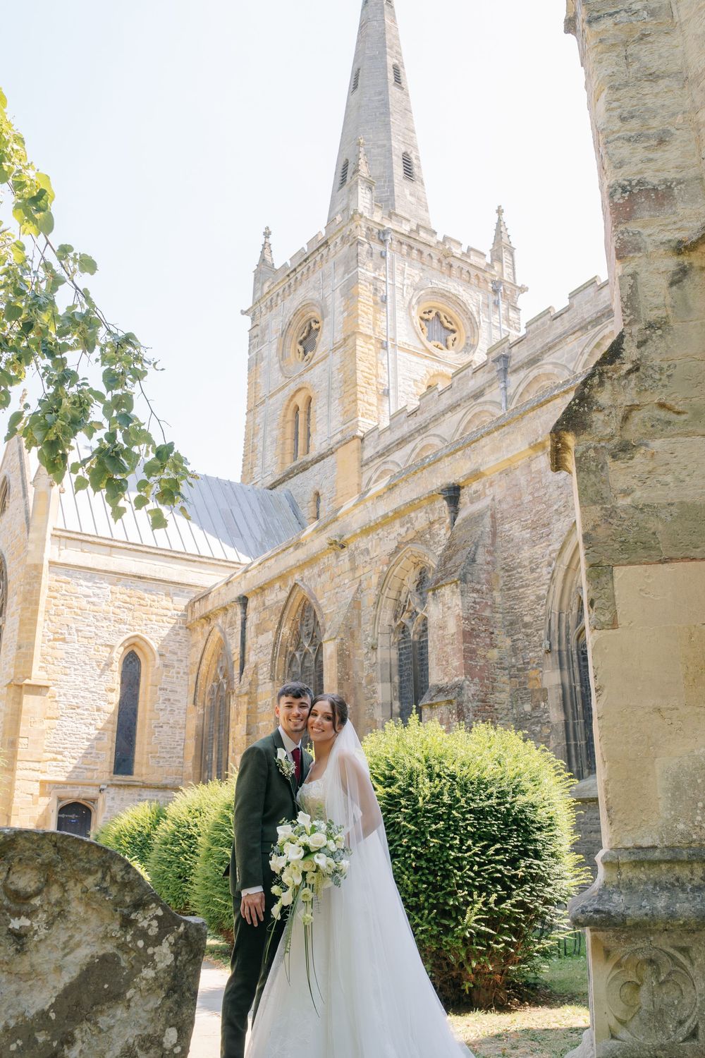Bride and groom pose together in front of a historic Gothic church with a tall spire on their wedding day. warwickshire wedding photographer