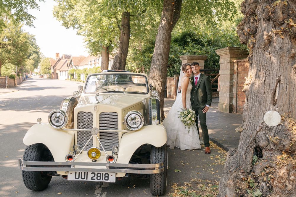 Bride and groom posing beside a vintage cream wedding car on a tree-lined street on their wedding day.