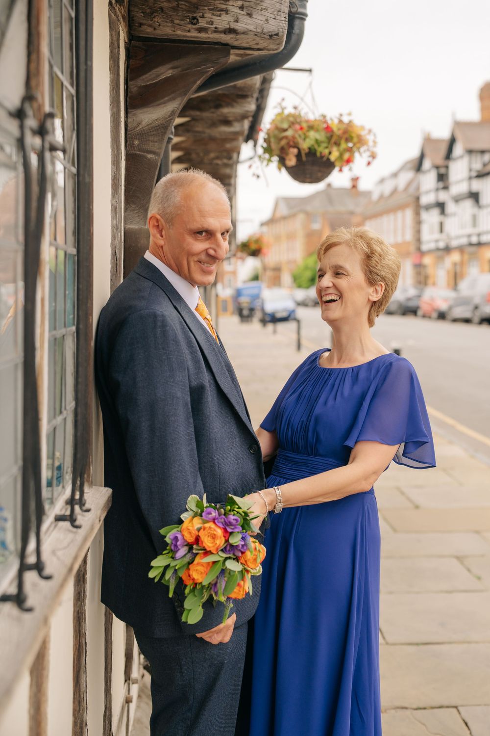 Mature couple in wedding attire laughing together on a town street, woman in blue dress holding orange bouquet.